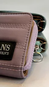 Billetera corta de diseño capitoneado con cierre perimetral y solapa frontal. Cuenta con compartimentos internos para tarjetas y billetes, e incluye una correa de mano desmontable.