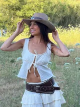 Top corto de color blanco con textura sutil, diseño sin mangas y escote redondo. Presenta detalles de encaje en los bordes y cierre frontal mediante lazos ajustables. El corte es holgado y ligero.