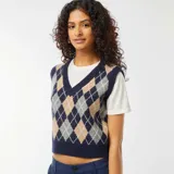 Chaleco de punto sin mangas, con cuello en V, de estilo argyle en tonos azul marino, beige, gris y blanco. Se muestra sobre una camiseta blanca de manga corta y un pantalón azul marino.