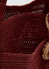 Bolso tote de tejido de fibra natural color bordó, con dos asas largas para hombro y detalle de charm colgante con flecos.