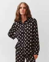 Camisa de corte recto y silueta fluida, color negro con estampado de lunares blancos. Presenta cuello clásico, cierre frontal de botones y canesú curvo sobre el pecho. Las mangas son largas, ligeramente abullonadas, con puños anchos abotonados y bajo redondeado.