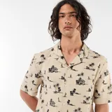 Camisa de manga corta color beige con estampado tropical de palmeras y veleros, cuello abierto y bolsillo en el pecho.