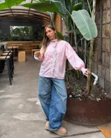 Camisa de manga larga abullonada en color rosa con rayas verticales finas. Presenta un diseño con volados en el cuello y a lo largo de la botonadura frontal, puños con detalle de rayas contrastantes y botones decorativos dorados.