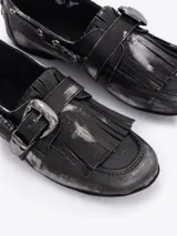 Zapato tipo mocasín de cuero vacuno con efecto gastado y detalles en color rojizo. Presenta flecos en la capellada, una hebilla metálica decorativa y herrajes plateados a lo largo del lateral.