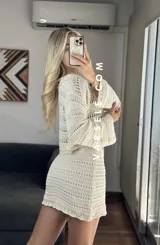 Conjunto de dos piezas tejido al crochet color crudo. Incluye una mini falda de tiro alto y un cardigan corto con mangas largas y lazos para atar en el frente.