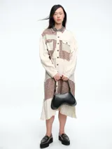 Vestido camisero de corte midi con diseño patchwork que combina paneles en tonos crudo y marrón con estampados florales sutiles. Presenta cierre frontal con botones, cuello camisero y mangas largas con puños.