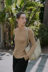 Bolso tipo shopper de hombro en color beige, con diseño estructurado, doble asa y detalle de pespuntes verticales en el cuerpo. Incluye un colgante decorativo tipo etiqueta y cierre imantado.