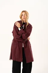 Trench de corte holgado color bordó, con cuello camisero, cierre frontal con botones y cinturón ajustable con hebilla. Presenta mangas largas con detalles de correas en los puños y bolsillos laterales.