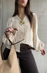 Camisa beige de manga larga con diseño abullonado y puños elastizados, escote en V con botones frontales y sutiles bordados florales tono sobre tono.
