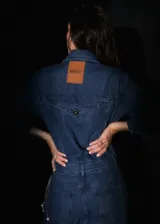Mono de denim azul con cierre frontal de botones metálicos, cuello camisero, mangas largas y bolsillos delanteros.
