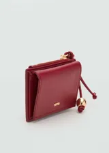 Monedero de efecto piel color rojo burdeos, con tamaño pequeño, cierre de solapa con botón magnético, bolsillo lateral con cremallera y detalle de abalorio colgante.
