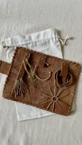 Set de accesorios compuesto por un brazalete rígido abierto, un collar con dije de sol radiante, un par de caravanas onduladas y un par de caravanas tipo fleco, presentados en un sobre de cuero con costuras artesanales.