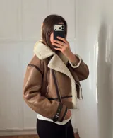 Campera tipo gamulán de cuero sintético en color tostado, con interior y solapas de corderito sintético en color crema. Presenta cierre frontal cruzado, bolsillos laterales con cierre y detalles de correas con hebillas en los puños.