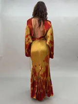 Vestido largo plisado con estampado abstracto en tonos rojo, naranja y dorado, cuello alto y mangas acampanadas. Incluye lazo fino para ajustar en la cintura.