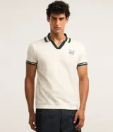 Remera polo comfort de algodón color marfil, con cuello en V verde oscuro con rayas blancas, mangas cortas con puños rayados y estampa de escudo bordada en el pecho.