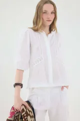 Camisa de lino color beige, de silueta romántica con escote a la base y mini volado. Cuenta con mangas 3/4 abullonadas con frunces y terminación en elástico. Posee detalles de puntillas negras en contraste en el frente y cierre frontal con botones ocultos bajo cartera.