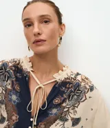 Blusa tipo bata de viscosa con estampa floral estilo paisley. Presenta cuello alto con detalle de volados, cierre frontal con botones y lazo ajustable en el escote. Posee mangas largas abullonadas con puños elásticos.