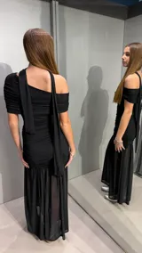 Vestido largo negro ajustado con diseño drapeado en todo el cuerpo, un hombro descubierto, cuello tipo choker integrado y falda larga semitransparente con vuelo en el inferior.