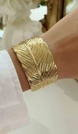 Brazalete ajustable de acero dorado con diseño en relieve en forma de hoja o pluma.