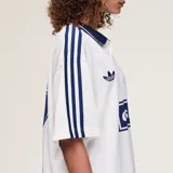 Camiseta Adidas Originals x Oasis de manga corta, color blanco con detalles en azul marino. Presenta cuello polo, las tres tiras características de la marca en los hombros, el logo del trifolio y un estampado frontal con la inscripción "oasis".