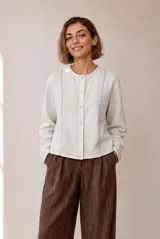 Chaquetita corta de lino color beige, con cuello redondo, manga larga y cierre frontal de botones metálicos a presión. Presenta costuras pespunteadas decorativas en la parte delantera.