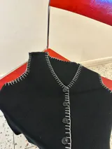Chaleco tejido color negro con corte sastrero y abotonadura frontal. Presenta un bordado a mano en hilo blanco en todo el contorno de la prenda, incluyendo cuello, sisas y borde inferior.