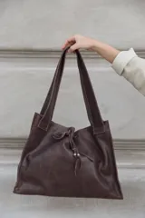 Bolso tipo shopper de cuero color marrón chocolate, con diseño amplio y cierre de cordón ajustable en la parte frontal. Cuenta con doble asa de hombro, forro interior, bolsillo interno y externo, y llavero incorporado.