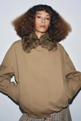 Chaqueta tipo bomber de color beige, con cuello solapa y aplicación extraíble de pelo sintético. Presenta cierre frontal con botones a presión ocultos, bolsillos delanteros y bajo con elástico.