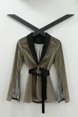 Blazer tipo smoking de terciopelo negro con acabado brillante y detalles estampados en dorado. Presenta solapas clásicas, cierre frontal cruzado mediante un lazo y mangas largas con cierres incorporados. El diseño es entallado y sofisticado.