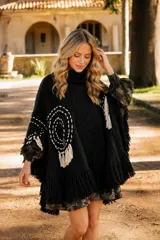Poncho de lana merino color negro con cuello alto, diseño amplio y flecos en el ruedo. Presenta detalles bordados circulares en color crudo y apliques de flecos decorativos en los laterales.