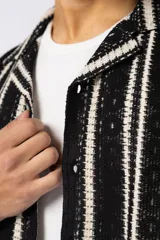 Camisa de manga corta con cuello clásico y cierre frontal de botones. Confeccionada en tejido de punto crochet calado, presenta un patrón de rayas verticales negras sobre fondo crudo.