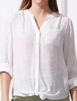 Blusa blanca de gasa marca Zac & Rachel, con cuello a la base, escote en V, abertura central con botones, bolsillo aplicado en el pecho y mangas largas regulables con presilla y botón. Tiene terminación opcional de nudo en el ruedo.