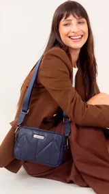 Bandolera de diseño rectangular con costuras geométricas acolchadas, doble compartimento con cierre metálico y correa de hombro ajustable en cinta técnica. Incluye placa metálica decorativa con logo en el frente.