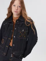 Campera denim crop de mezclilla oscura, con apliques metálicos en el pecho, bolsillos delanteros cruzados con detalles bordados y de parches.