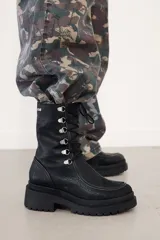 Borcego de caña corta confeccionado en cuero negro, con diseño de cordones y ganchos metálicos, cierre lateral y suela de plataforma dentada.