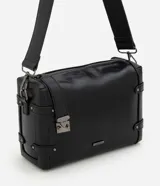 Cartera tipo shoulder mediana de formato rectangular, confeccionada en poliuretano color negro con efecto texturizado. Presenta un diseño moderno con herrajes metálicos oscuros, cierre superior con cremallera, correas laterales decorativas con tachas y una correa ancha ajustable para el hombro.