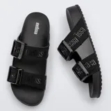 Sandalias Melissa Cozy Slide M-Lover color negro, con dos tiras anchas con hebillas grandes y apliques con logo. Suela de PVC extra flexible.