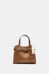 Bolso tipo mini shopper de cuero sintético color marrón caramelo, con costuras blancas visibles. Posee asas de mano y una correa bandolera ancha de tela color beige. El frente tiene un detalle de tira con hebilla metálica dorada.