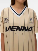 Camiseta de estilo deportivo color beige con rayas verticales negras, cuello en V con detalles en blanco y negro, y estampado frontal con la palabra VIENNE.