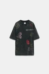 Camiseta oversize de algodón con efecto lavado, cuello redondo y manga corta. Presenta múltiples aplicaciones de brillos y strass con motivos coloridos de Smiley, incluyendo estrellas, corazones y caras sonrientes.