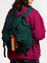 Mochila acolchada de nylon color verde oscuro con detalles en negro. Presenta un compartimento principal con cierre de cordón y tapa con hebilla de plástico negra. Incluye un bolsillo frontal con tapa y hebilla. Posee tiras ajustables negras y un tirador naranja con la marca "Rusty" visible.