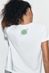 Camiseta de algodón blanca con cuello redondo y manga corta. Presenta un estampado gráfico frontal con dibujos estilo garabato y texto en tonos rojizos.