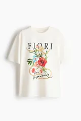 Remera oversized de algodón color blanco, con cuello redondo, mangas cortas y hombros caídos. Presenta un estampado frontal con la palabra 'Fiori', ilustraciones de flores coloridas y cadenas doradas.