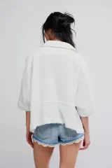 Blusa blanca de algodón vaporoso, con diseño holgado y ligeramente extragrande, cuello camisero, cierre frontal de botones y mangas amplias.