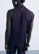 Blusa sin mangas de corte recto con cuello drapeado y cierre de botones. Presenta un diseño estampado de lunares en color azul sobre fondo oscuro.