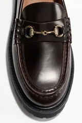 Mocasines de tejido revestido en color marrón oscuro, con diseño tradicional, detalle de estribo metálico dorado en el empeine y suela gruesa tractorada de color negro.
