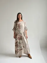 Vestido largo beige de lino con diseño midi holgado, cuello redondo, mangas cortas con vuelta y un gran estampado lateral de flores y hojas en tonos marrones y verdes. Cuenta con un bolsillo frontal aplicado con detalles deshilachados.