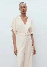 Mono largo de corte recto y color beige, con cuello en pico, mangas cortas y cinturón incorporado con hebilla metálica. Cuenta con cierre de cremallera en la parte posterior.