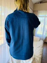 Sobrecamisa de gamuza con cuello clásico, cierre frontal con botones metálicos y mangas largas con puños abotonados. Presenta un diseño estructurado con corte recto y largo a la cadera.