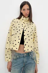 Chaqueta de cuello subido y manga larga con estampado de lunares negros sobre base beige. Cuenta con bolsillos de vivo en los laterales, detalles de frunces ajustables con cordones en el delantero y cierre frontal con botones.